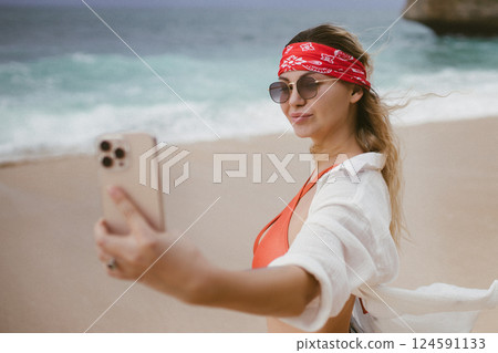 Beautiful happy woman doing selfie on beach summer. Young smiling girl making selfie on smartphone over sea background, cloudy weather. Romantic lady with laughing emotional face make photo on phone 124591133