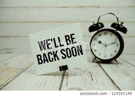We'll Be Back Soon text on paper card with alarm clock on wooden background 124591149