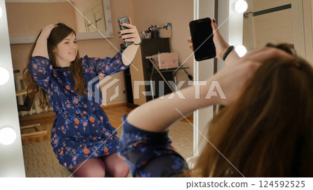 The girl sits on the floor and makes selfies at the makeup mirrors at home. The girl sits on the floor and makes selfies at the makeup mirrors at home. 124592525