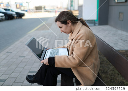 Woman focused on laptop while sitting on bench in urban area during daytime 124592529