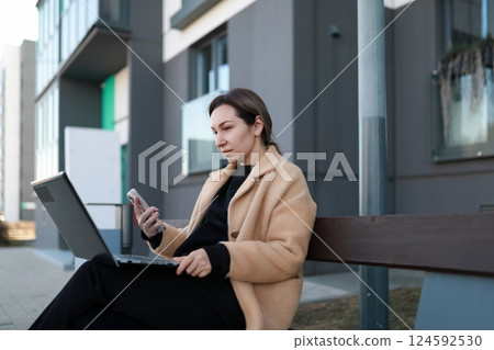 Young woman using laptop and smartphone while sitting on a bench outside a modern building in the Young woman using laptop and smartphone while sitting on a bench outside a modern building in the 124592530