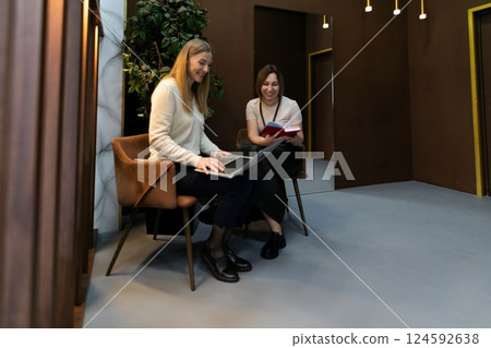 Two women collaborate in a modern workspace while using a laptop and a book in an indoor setting 124592638