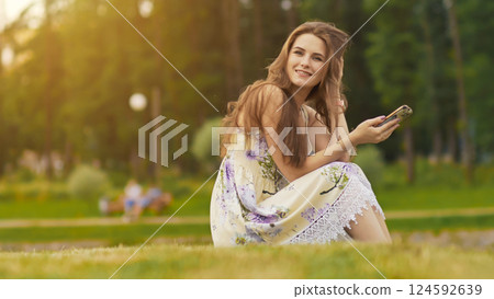 Young beautiful woman in summer dress with long hair sitting on grass in green park and talking on the phone, smiling. Summer. Recreation. Youth. 124592639