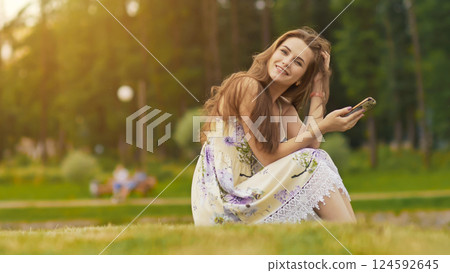 Young beautiful woman in summer dress with long hair sitting on grass in green park and talking on the phone, smiling. Summer. Recreation. Youth. 124592645