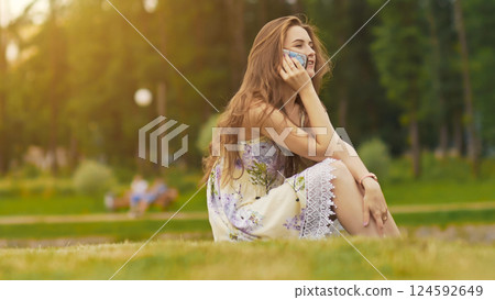 Young beautiful woman in summer dress with long hair sitting on grass in green park and talking on the phone, smiling. Summer. Recreation. Youth. 124592649