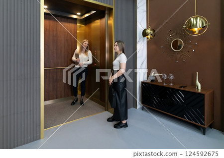 Two women interact in a modern elevator while dressed stylishly in a chic interior setting during 124592675