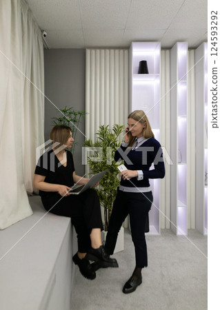 Two women engaged in conversation while working in a modern office space with plants and stylish Two women engaged in conversation while working in a modern office space with plants and stylish 124593292