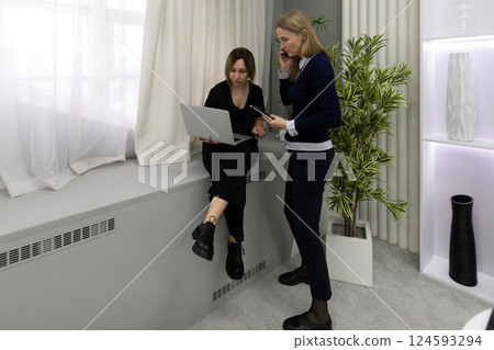 Two women discussing ideas while working with a laptop and phone in a modern office space during 124593294