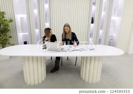 Two women collaborating at a minimalist workspace with documents and a laptop in a modern office 124593353