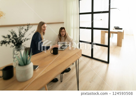 Two women collaborating at a wooden table in a bright office setting while using a laptop for a work 124593508