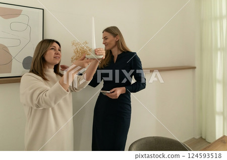 Two women decorating a modern interior space with a candle and floral arrangement during daytime 124593518