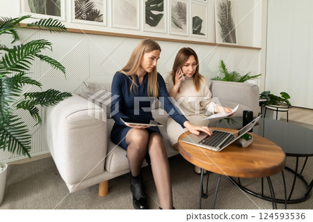 Two women collaborating on a laptop while sitting on a sofa in a modern, plant-filled office space 124593536