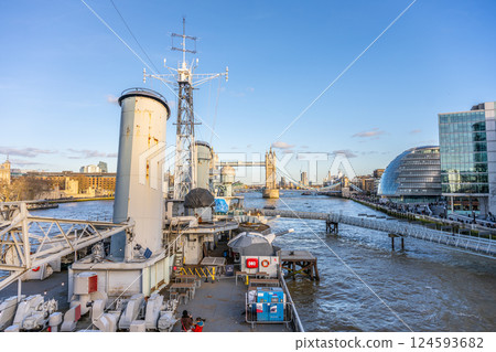 Visitors explore the deck and funnels of HMS Belfast while taking in the stunning views of the River Thames and Tower Bridge in London, showcasing the city's rich maritime history. 124593682