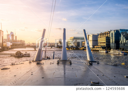 Turrets of the HMS Belfast rise prominently against the skyline of London. The sun sets behind modern buildings, casting warm light over the River Thames and nearby boats. 124593686