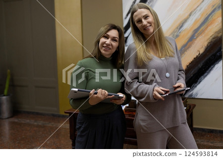 Two women smiling and holding tablets while standing next to an abstract painting in a modern indoor Two women smiling and holding tablets while standing next to an abstract painting in a modern indoor 124593814