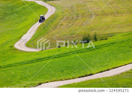 Scenic aerial view small tractor drive winding dirt path road green fields at bright summer sunshine day. Farmland slopes countryside tranquil exploration. Panoramic alpine agriculture machine scene Scenic aerial view small tractor drive winding dirt path road green fields at bright summer sunshine day. Farmland slopes countryside tranquil exploration. Panoramic alpine agriculture machine scene 124595271