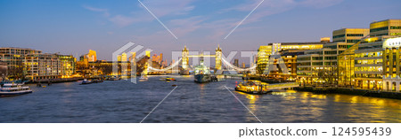 Tower Bridge and HMS Belfast stand majestically over the Thames River during a tranquil evening in London, illuminating the skyline with warm lights as day turns to night. 124595439