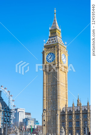 Big Ben tower with its detailed clock face and golden facade stands tall beside the Gothic Palace of Westminster, with the London Eye partially visible. 124595440