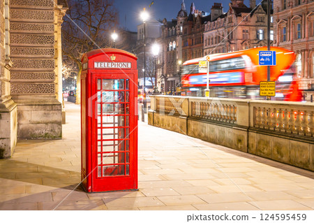 A classic red telephone box stands illuminated on a quiet street in London. Night falls as buses pass by, highlighting the city's vibrant nocturnal atmosphere. 124595459