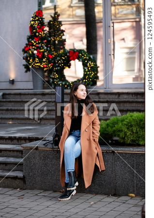 Young woman enjoys a quiet moment on a stone bench in front of festive holiday decorations in a city park on a crisp winter afternoon 124595493