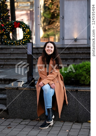 Cheerful woman enjoying the festive atmosphere while relaxing on a city bench adorned with holiday decorations at dusk 124595494