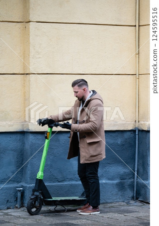 Man in a brown coat checking a green electric scooter against a textured wall in an urban setting during a chilly afternoon Man in a brown coat checking a green electric scooter against a textured wall in an urban setting during a chilly afternoon 124595516