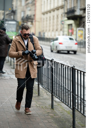 Walking through a bustling city street dressed in a stylish coat while capturing urban moments with a camera in hand during a chilly afternoon Walking through a bustling city street dressed in a stylish coat while capturing urban moments with a camera in hand during a chilly afternoon 124595520