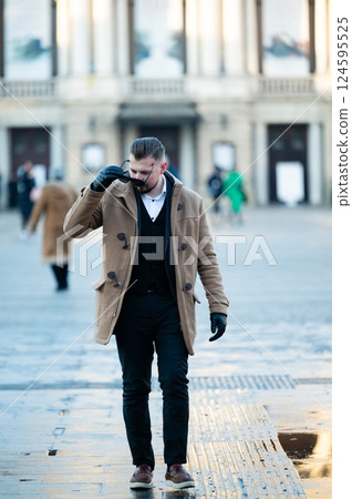 Elegant man strolls casually through a snowy plaza, adjusting his stylish scarf while the winter sun illuminates the historic architecture in the background Elegant man strolls casually through a snowy plaza, adjusting his stylish scarf while the winter sun illuminates the historic architecture in the background 124595525