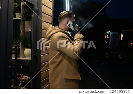 Warm coat, cool night phone call amidst smoky ambiance and urban greenery near illuminated shops enhancing the evening atmosphere Warm coat, cool night phone call amidst smoky ambiance and urban greenery near illuminated shops enhancing the evening atmosphere 124595535