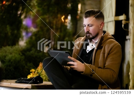 Man engrossed in tablet while seated outside among lush greenery in a cozy evening setting illuminated by soft lights 124595539