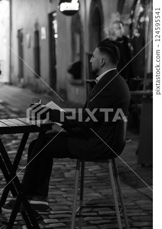 Lonely figure enjoying a quiet evening in a cobblestone alley while sipping a drink and reading a book in a charming old town setting 124595541