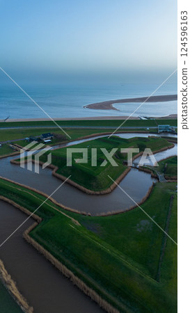 A drone video showing a unique landscape of man-made mounds and waterways near the sea, with a house on the grassy terrain, and a distant view of the coastline stretching under a cloudy sky. A drone video showing a unique landscape of man-made mounds and waterways near the sea, with a house on the grassy terrain, and a distant view of the coastline stretching under a cloudy sky. 124596163