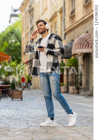 Overjoyed Arabian man in wireless headphones listening energetic music on smartphone in city street 124596302