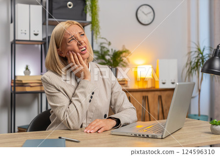 Upset mature businesswoman in suit suffering from toothache while using laptop at home office desk 124596310