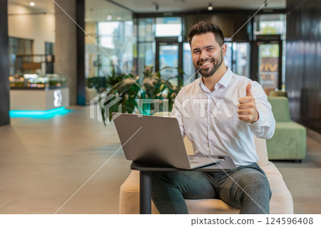 Happy young businessman working on laptop looking at camera showing thumbs up in modern office lobby Happy young businessman working on laptop looking at camera showing thumbs up in modern office lobby 124596408