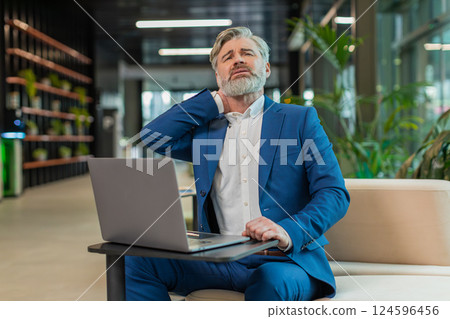 Tired middle-aged businessman suffers from spine neck muscle pain working on laptop in office lobby Tired middle-aged businessman suffers from spine neck muscle pain working on laptop in office lobby 124596456