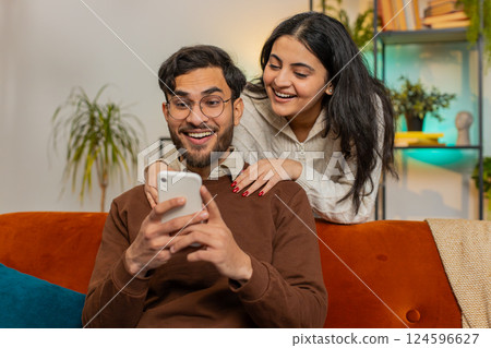 Smiling Indian woman talking with boyfriend swiping smartphone sitting on sofa at home family couple Smiling Indian woman talking with boyfriend swiping smartphone sitting on sofa at home family couple 124596627