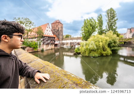 Nuremberg, Germany, August 1, 2023. A young Caucasian man is fascinated by the enchanting view of the river with the half-timbered house reflected in the water. Exploring and discovering Nuremberg. Nuremberg, Germany, August 1, 2023. A young Caucasian man is fascinated by the enchanting view of the river with the half-timbered house reflected in the water. Exploring and discovering Nuremberg. 124596713
