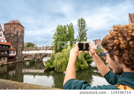 Nuremberg,Germany, August 1, 2023. A young Caucasian man is fascinated by the enchanting view of the river with the half-timbered house reflected in the water. He uses his cell phone to photograph it. Nuremberg,Germany, August 1, 2023. A young Caucasian man is fascinated by the enchanting view of the river with the half-timbered house reflected in the water. He uses his cell phone to photograph it. 124596714