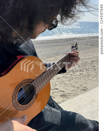 Long Hair Boy Plays Classical Guitar by the Ocean in Summer Dressed in Black 124597840