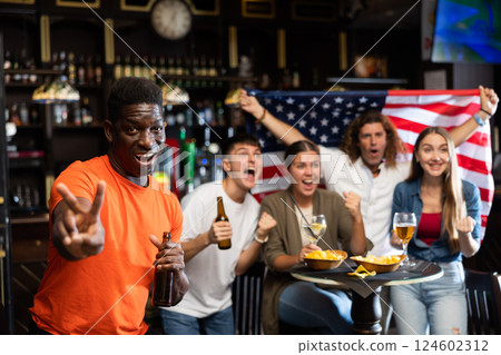 Happy multiracial sport fans holding the flag of USA, enjoing game, beer and chips in the bar 124602312