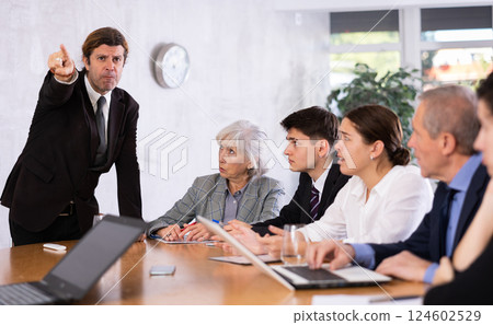 Furious male boss yelling at office colleagues of different ages taking notes at table with laptops Furious male boss yelling at office colleagues of different ages taking notes at table with laptops 124602529