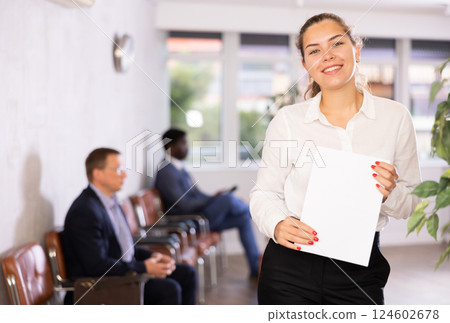 Friendly hostess woman stands at office reception with list of guests and expects invited visitors Friendly hostess woman stands at office reception with list of guests and expects invited visitors 124602678