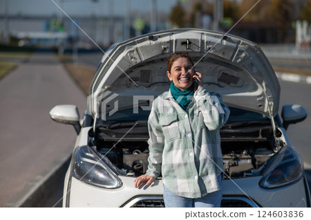 Caucasian woman laughing and calling on phone asking for help standing near car with open hood.  124603836