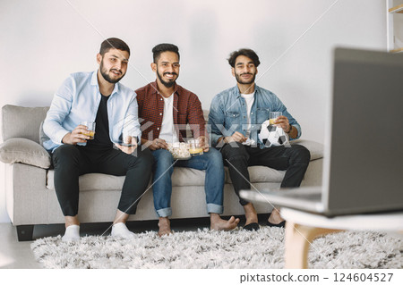 Three young excited male friends sitting on a coach while watching football match on laptop. Men drinking juice and eating popcorn. Men wearing shirts and t-shirts. 124604527