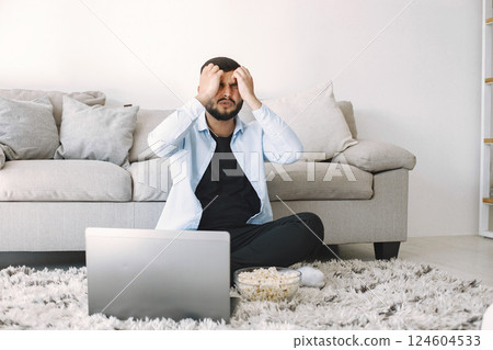 Young disappointed bearded man sitting on a floor near the couch while watching football match on laptop. Man holding his hands on his head. Man in blue shirt sitting on carpet. Young disappointed bearded man sitting on a floor near the couch while watching football match on laptop. Man holding his hands on his head. Man in blue shirt sitting on carpet. 124604533