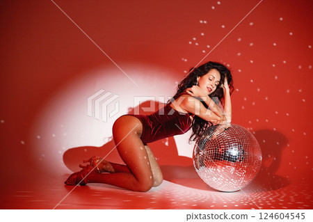 Curly brunette girl sitting near big disco ball and posing for a photo. Girl wearing velour bodysuit. Projectors light girl up. 124604545