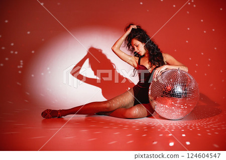 Curly brunette girl sitting near big disco ball and posing for a photo. Girl wearing velour bodysuit. Projectors light girl up. 124604547
