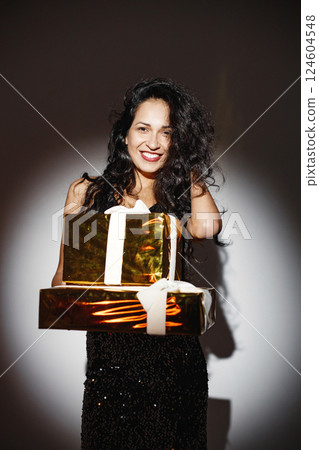 Woman holding a wrapped in golden paper gift boxes in hands. Curly brunette girl standing near white wall and posing for a photo. Girl wearing black skirt and top. 124604548