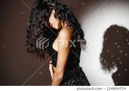 Curly brunette girl standing near white wall and posing for a photo. Girl wearing black skirt and top. Projectors light girl up. Curly brunette girl standing near white wall and posing for a photo. Girl wearing black skirt and top. Projectors light girl up. 124604554